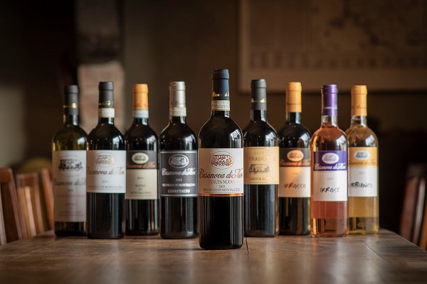 Auf einem Holztisch stehen zehn Flaschen Wein in verschiedenen Farben und Etiketten. Die zentrale Flasche ist eine rote, während die anderen aus Weiß- und Roséweinen bestehen, wobei sie in einer attraktiven Anordnung präsentiert sind.