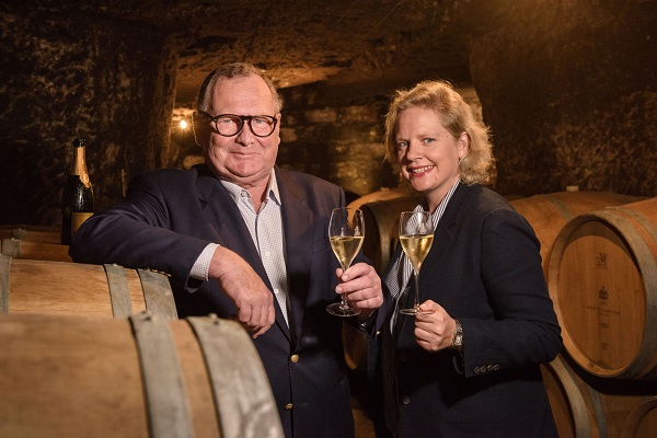 Zwei Personen stehen in einem Weinkeller. Sie halten Gläser mit Sekt und lächeln. Im Hintergrund sind Holzfässer und schwaches, warmes Licht sichtbar, was eine gemütliche Atmosphäre schafft.