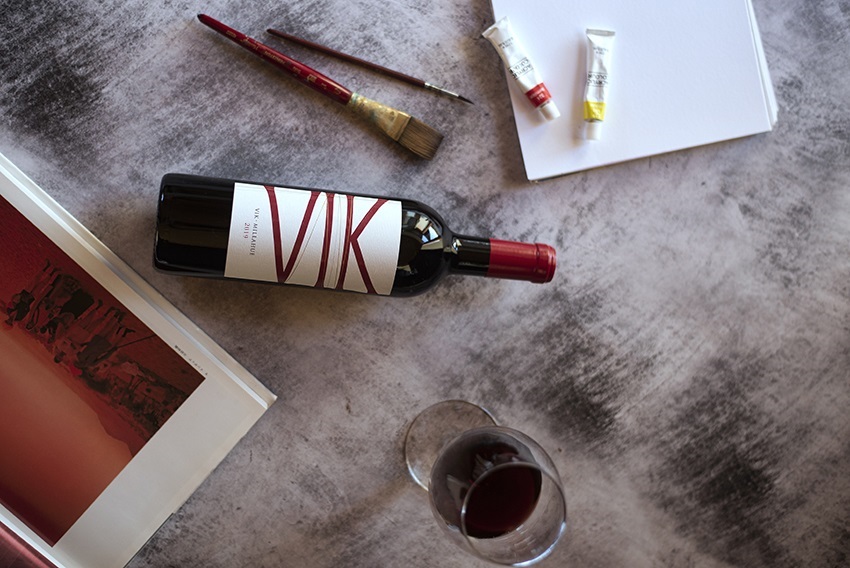 Eine Weinflasche mit einem eleganten Etikett liegt auf einem Tisch neben einem Glas Rotwein, Pinsel, zwei Farbtuben und einem Block Papier, umgeben von einem Stoff.