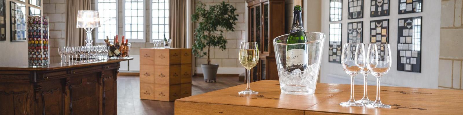 Ein eleganter Raum mit einem Holzbar, auf dem eine Flasche Sekt in einem Eiskübel steht. Neben dem Kübel befinden sich mehrere Weingläser. Im Hintergrund sind eine Pflanze und Galeriewände zu sehen.