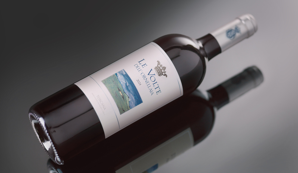 Eine Weinflasche liegt schräg auf einer reflektierenden Oberfläche. Das Etikett zeigt den Namen -Le Volte dell-Ornellaia- aus dem Jahr 2014 und eine Landschaftsillustration, umgeben von schlichten Farben.