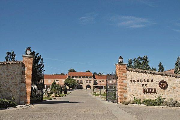 Eine große Zufahrt führt durch ein offenes Tor zu einem eleganten Gebäude im Hintergrund. Umgeben von Bäumen und einem klaren blauen Himmel. Die Einfahrt ist gepflastert.