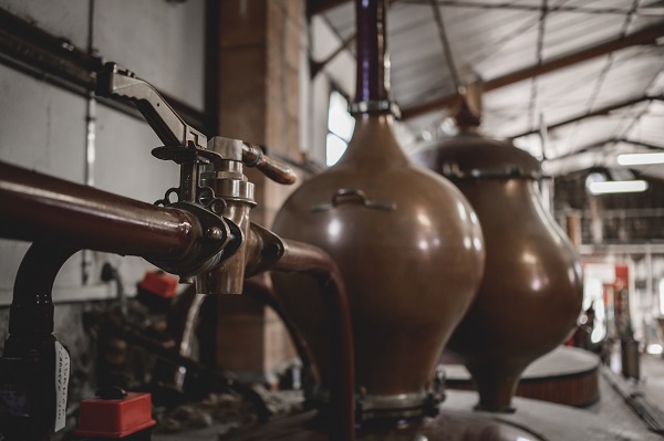 Eine kupferne Destillationsvorrichtung steht im Vordergrund, während im Hintergrund weitere große kupferne Kessel zu sehen sind. Der Raum wirkt industriell und gut beleuchtet.