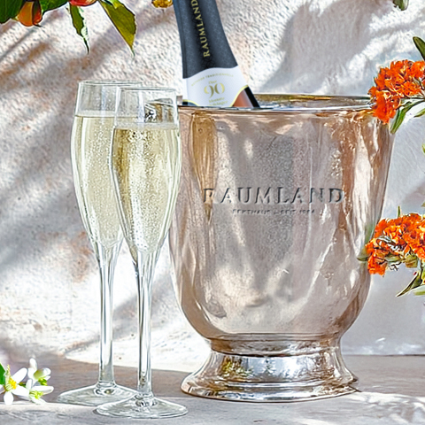 Zwei stilvolle Sektgläser stehen neben einem eleganten, silbernen Sektkübel, aus dem eine Flasche herausragt. Umgeben sind sie von frischen Blumen, die eine festliche Atmosphäre schaffen.