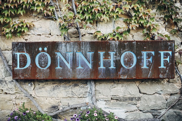 Ein rostiges Schild mit der Aufschrift -DÖNNHOFF- steht an einer Steinmauer, die von Efeu und bunten Blumen umgeben ist. Vines wachsen über das Schild und die Mauer.