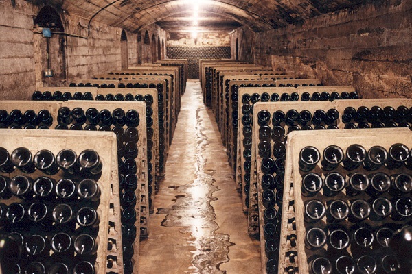 Eine Weinkeller-Umgebung mit Reihen von gestapelten Weinflaschen auf Regalen. Der Raum hat eine gewölbte Decke und ist schwach beleuchtet, mit einem fließenden Wasserweg auf dem Boden.