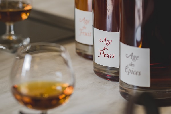 Drei Flaschen mit Etiketten stehen auf einem Tisch. Die Etiketten tragen die Aufschriften -Age des Fleurs-, -Age des Fruits- und -Age des Épices-. Im Vordergrund steht ein Glas mit aromatischem Getränk.