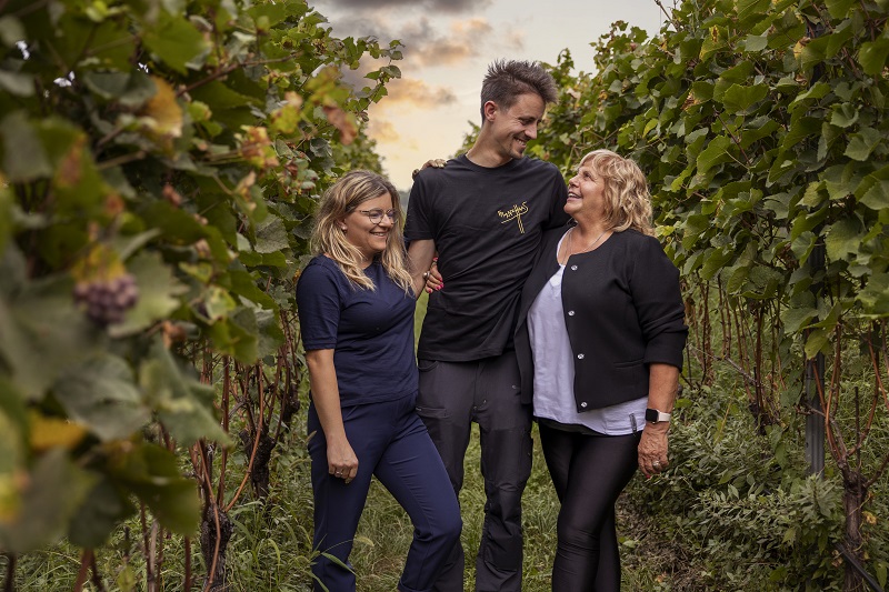 Drei Personen stehen zusammen in einem Weinberg, umgeben von Weinstöcken. Sie lächeln und scheinen eine fröhliche Unterhaltung zu führen, während die Sonne im Hintergrund untergeht.
