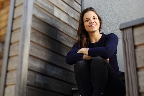 Eine Frau sitzt fröhlich auf einer Treppe, die auf eine Holzverkleidung führt. Sie trägt einen dunkelblauen Pullover und hat lange, lockige Haare. Der Hintergrund ist einfach und hell.