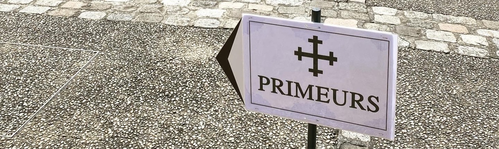 Ein Schild mit der Aufschrift -PRIMEURS- zeigt in Richtung einer Straße. Der Boden besteht aus grauem, grobem Asphalt mit gelegentlichen Kopfsteinpflastersteinen.