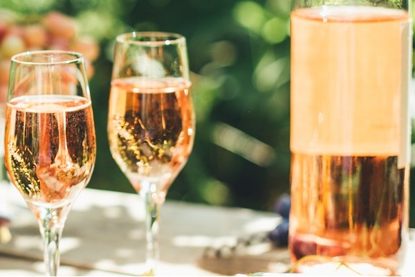 Zwei Gläser mit roséfarbenem Sekt stehen auf einem Tisch, neben einer großen Flasche des gleichen Getränks. Im Hintergrund sind unscharfe grüne und rote Elemente sichtbar.