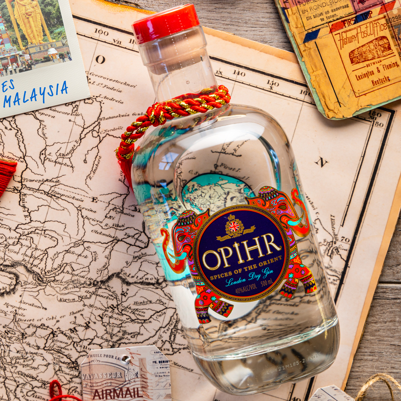 Eine Flasche OPIHR London Dry Gin steht auf einer alten Karte, umgeben von Postkarten und Airmail-Etiketten. Die Flasche hat einen roten Deckel und ist mit einem bunten Elefantenmuster verziert.
