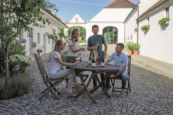 Vier Personen sitzen an einem Tisch im Freien, genießen Getränke und unterhalten sich. Die Umgebung zeigt ein gepflastertes Gelände mit Pflanzen und einem hellen, sonnigen Hintergrund.
