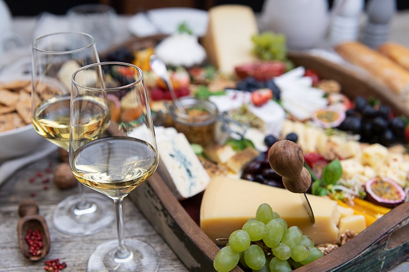 Eine elegante Käseplatte mit verschiedenen Käsesorten, Trauben, Nüssen und Marmelade ist liebevoll angerichtet. Zwei Gläser Weißwein stehen daneben, und im Hintergrund sind Brot und Geschirr sichtbar.