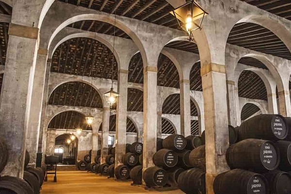 Fässer stehen in einem großen, architektonisch gestalteten Lagerraum. Der Raum hat hohe Gewölbedecken und wird von Laternen beleuchtet. Die Wände sind aus Beton oder Stein und wirken rustikal.
