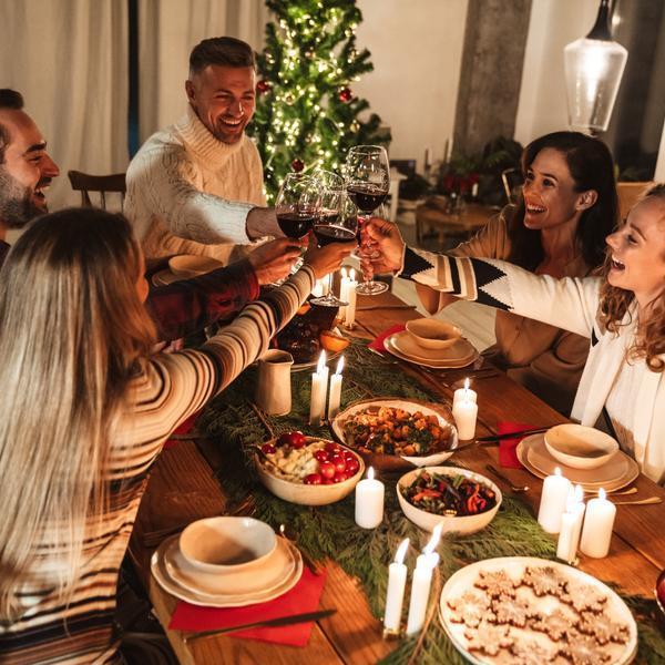 Sechs Personen heben ihre Weingläser zum Anstoßen an einem festlich gedeckten Tisch mit verschiedenen Gerichte und Kerzen. Im Hintergrund steht ein Weihnachtsbaum, der das Ambiente festlich gestaltet.