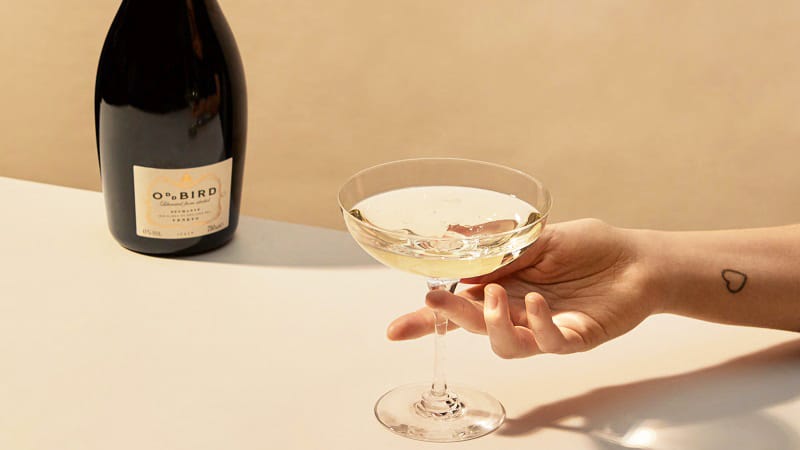 Ein Hand reicht ein elegantes Glas mit einem sprudelnden Getränk. Im Hintergrund steht eine Flasche O.BIRD. Die Oberfläche ist hell und minimalistisch.