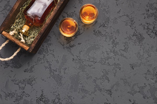 Eine transparente Flasche mit einer bernsteinfarbenen Flüssigkeit liegt in einer Holzkiste, die mit Heu ausgekleidet ist. Der Deckel der Kiste liegt daneben auf dem Tisch.