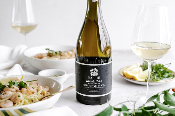 Eine Flasche Babich Sauvignon Blanc steht im Vordergrund. Daneben gibt es ein Glas Weißwein und eine Schüssel mit Pasta und Garnelen auf einem Tisch, dekoriert mit frischen Kräutern und Zitronenscheiben.