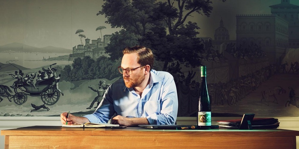 Ein Mann mit Brille sitzt an einem Holztisch und schreibt in ein Notizbuch. Er ist in einem hellblauen Hemd gekleidet. Im Hintergrund ist eine muralartige Wandgestaltung zu sehen, und auf dem Tisch steht eine Flasche.