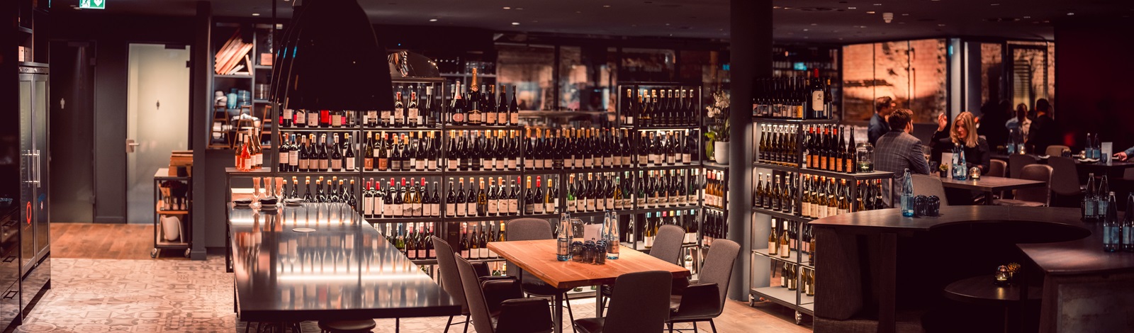 Ein elegantes Restaurant mit einem langen, glänzenden Tisch, umgeben von Stühlen. Im Hintergrund sind Weinregale voller Flaschen sichtbar, während Gäste an anderen Tischen sitzen und sich unterhalten.