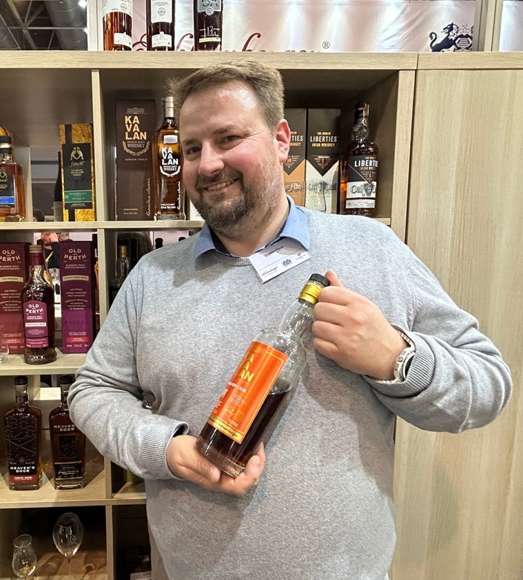 Ein Mann hält eine Flasche Whisky mit orangefarbenem Etikett. Im Hintergrund stehen verschiedene Whiskyflaschen in Regalen. Der Mann lächelt und trägt einen grauen Pullover.