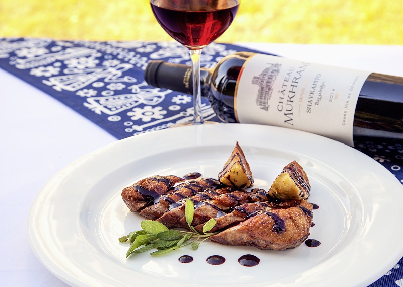 Gegrilltes Hähnchenfilet mit einer Glasur ist auf einem weißen Teller angerichtet, umgeben von Zwiebelstücken und Kräutern. Im Hintergrund steht ein Glas Rotwein neben einer Weinflasche auf einem dekorativen Tischläufer.