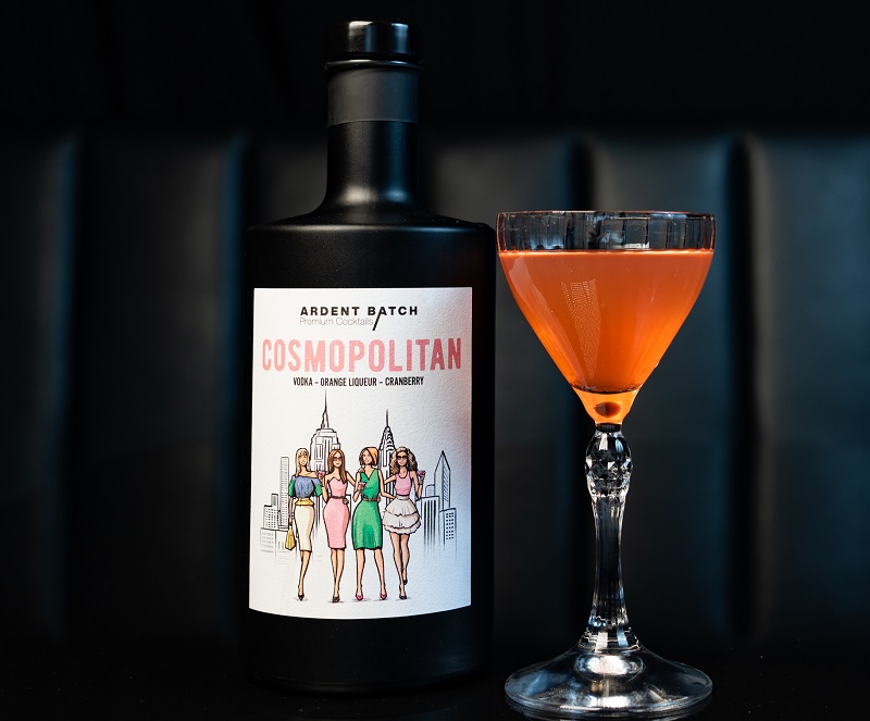 Eine Flasche Cosmopolitan-Cocktailmix mit einer stilisierten Abbildung von Frauen in Farben steht neben einem gefüllten Cocktailglas, das einen rosa-orangen Drink zeigt, vor einem dunklen Hintergrund.