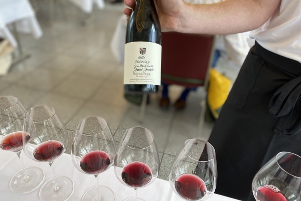 Ein Kellner hält eine Flasche Wein mit Etikett und schenkt roten Wein in mehrere Gläser ein, die auf einem Tisch stehen. Der Hintergrund zeigt unscharfe Tische im Restaurant.
