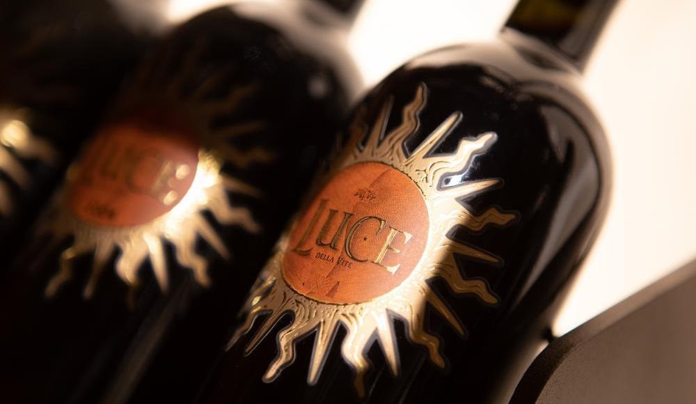 Drei Flaschen Wein mit einem goldenen Sonnen-Design und dem Wort -JUICE- in der Mitte. Sie stehen auf einem Regal und sind vor einem hellen Hintergrund positioniert.