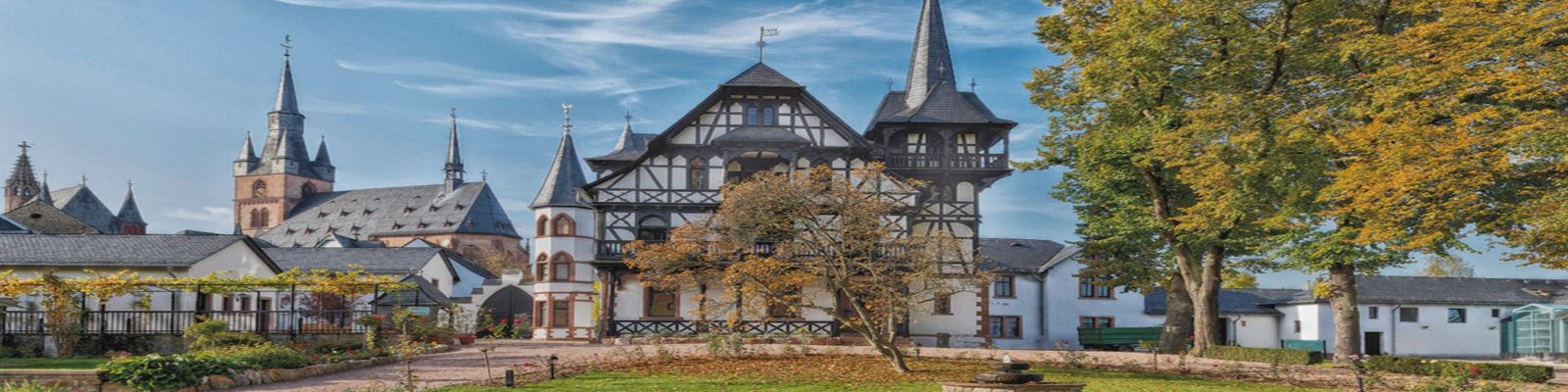 Ein historisches Gebäude mit Fachwerkarchitektur steht im Vordergrund, umgeben von Bäumen im Herbst. Im Hintergrund sind mehrere Kirchtürme und weitere Gebäude sichtbar, unter blauem Himmel.