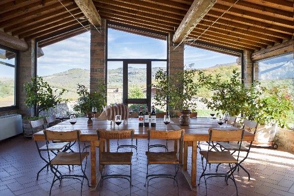 Ein großer Holztisch mit Stühlen steht in einem lichtdurchfluteten Raum. Große Fenster bieten einen Blick auf sanfte Hügel und Grünpflanzen. Der Raum hat eine rustikale Atmosphäre.