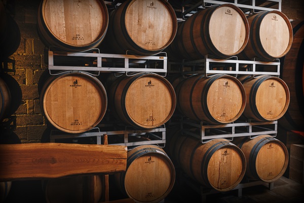 Eichenfässer stehen in mehreren Reihen auf einem Metallregal. Die Fässer sind aus hellem Holz und wirken robust. Im Hintergrund ist eine schummrige Kelleratmosphäre zu erkennen.