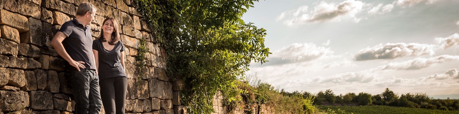 Christmann Ein Paar steht lächelnd an einer Steinmauer, umgeben von Bäumen und einer hügeligen Landschaft. Der Himmel ist bewölkt, und die Szenerie wirkt harmonisch und entspannt.