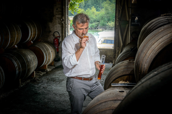Ein Mann steht in einem dunklen Raum zwischen großen Holzfässern. Er riecht an einem Glas und betrachtet aufmerksam seine Umgebung, während Tageslicht durch die Tür strömt.