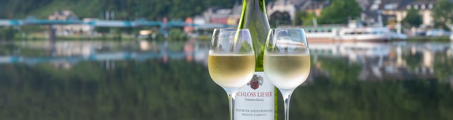 Eine Flasche Riesling und zwei Gläser mit Weißwein stehen am Ufer eines Flusses. Im Hintergrund sind verschwommene Gebäude und ein grüner Hügel zu sehen, reflektiert im Wasser.