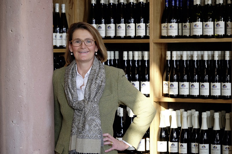Eine Frau in einem eleganten Outfit steht lächelnd neben einem Regal voller Weinflaschen. Der Hintergrund zeigt eine Wand aus Holz mit vielen Flaschen, die etikettiert sind.