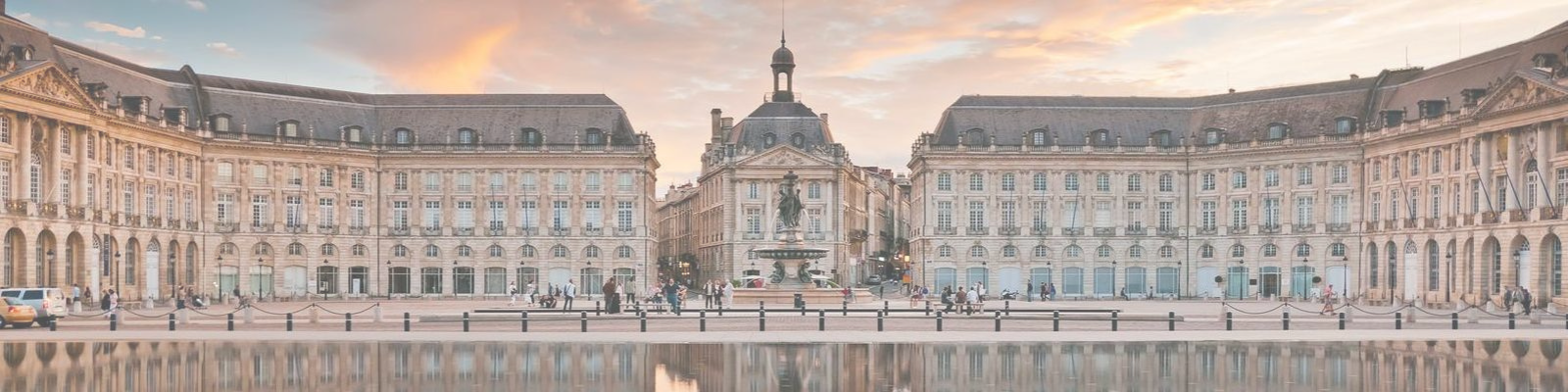 Eine große Platzanlage mit historischen Gebäuden umgeben, Menschen laufen herum. In der Mitte steht eine Statue, während sich die Wolken über dem Platz färben. Eine spiegelnde Wasserfläche reflektiert die Architektur.