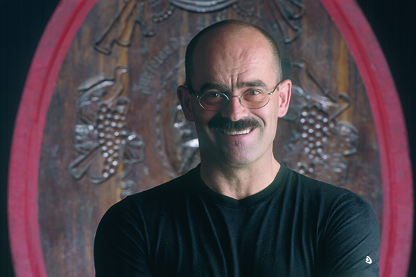 Ein bärtiger Mann mit Brille trägt ein schwarzes T-Shirt und lächelt. Er steht vor einem kunstvoll verzierten Holzschild mit Weintraubenmotiven im Hintergrund.