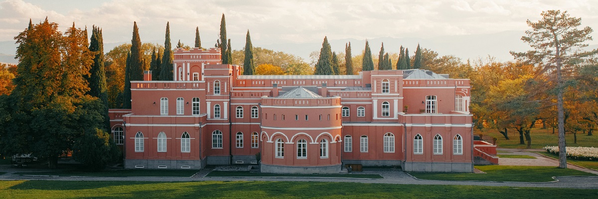 Ein farbenfrohes, historisches Gebäude mit rosa Fassade und weißen Fenstern steht in einem grünen Park. Umgeben von Bäumen und einem klaren Himmel, vermittelt es eine ruhige Atmosphäre.