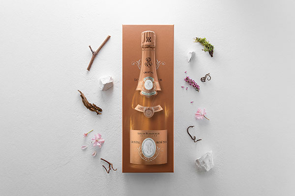 lr_cristal_rose_and_its_pack_with_a_floral_composition_louis_roederer_low_res_600x400 Ein eleganter Sekt in einer kunstvoll gestalteten Verpackung liegt auf einem hellen Untergrund. Um die Flasche sind verschiedene natürliche Elemente wie Zweige, Blüten und kleine Steine verteilt.