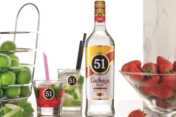 Eine Flasche Cachaca 51 steht im Mittelpunkt, umgeben von zwei Cocktails mit Limetten und Erdbeeren. Links sind grüne Limetten in einem Gestell und den Erdbeeren auf der rechten Seite.