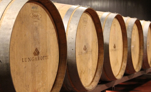 Holzfässer stehen aufgereiht in einem Raum, ihre Metallbänder glänzen leicht. Einige Fässer sind mit -Lungarotti- graviert, die Umgebung wirkt düster und rustikal.