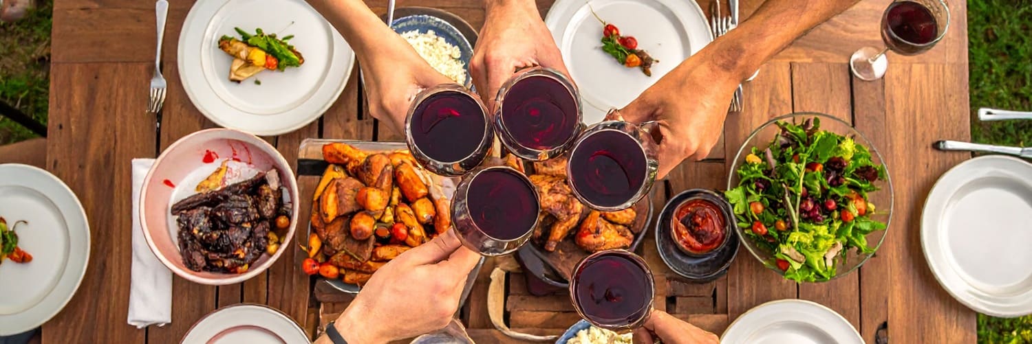 Sechs Hände heben Gläser mit Rotwein und einem Flaschenbier, um zu stoßen. Im Hintergrund sind verschiedene Gerichte wie Pasta und gegrillte Maiskolben auf einem Tisch zu sehen.