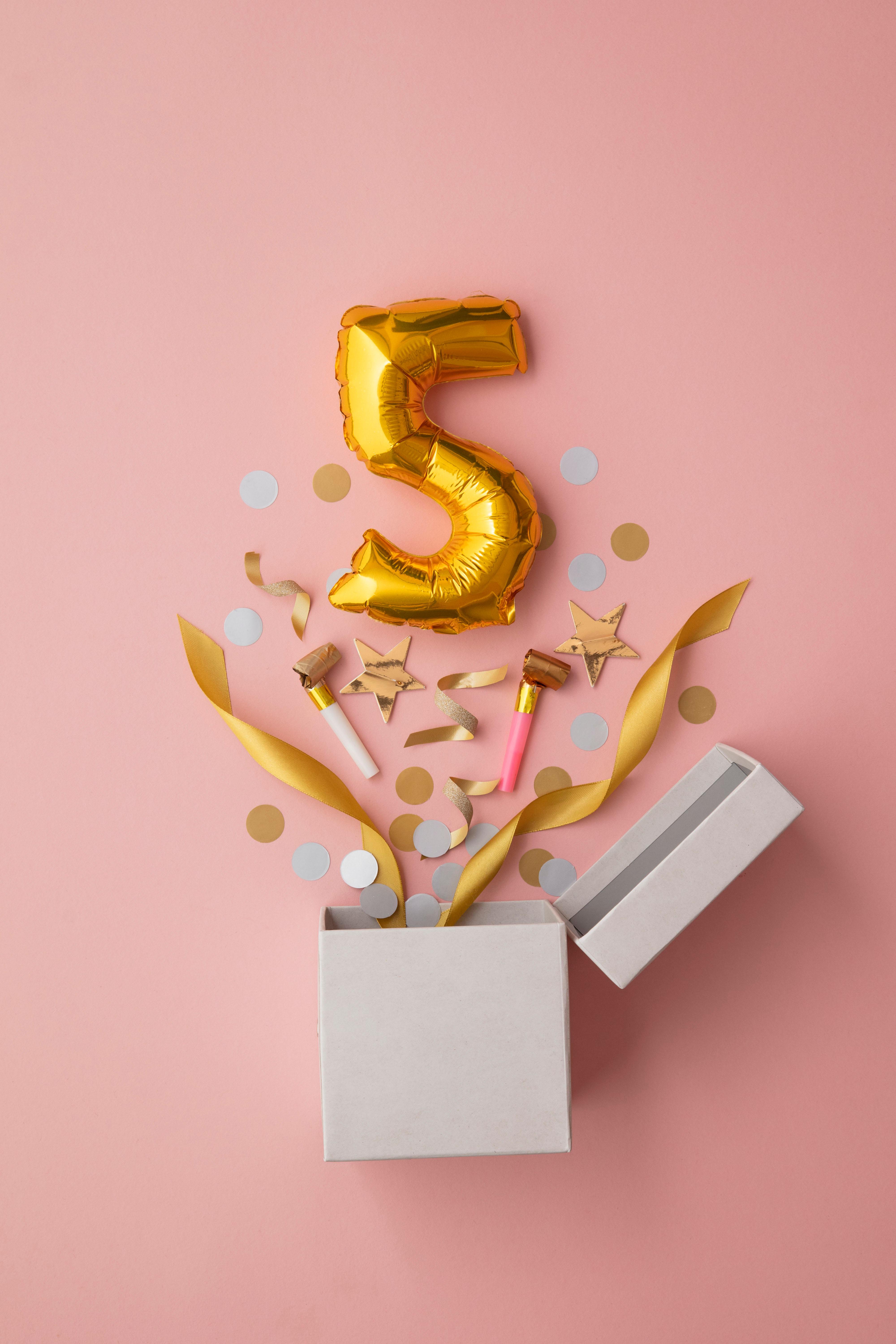 Ein offener Geschenkkarton entfaltet Konfetti, goldene Sterne, Luftschlangen und Knaller. Darüber schwebt eine goldene -5--Ballonzahl, alles vor einem rosa Hintergrund.