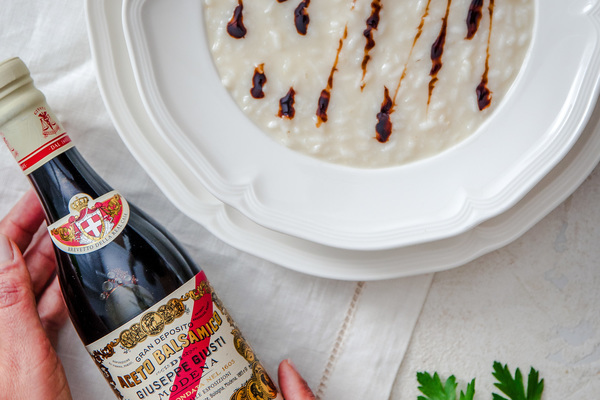 Eine Flasche Balsamico-Essig wird in einer Hand gehalten, während ein Teller mit cremigem Risotto im Hintergrund zu sehen ist. Das Risotto ist mit dunklen Balsamico-Drizzles garniert.