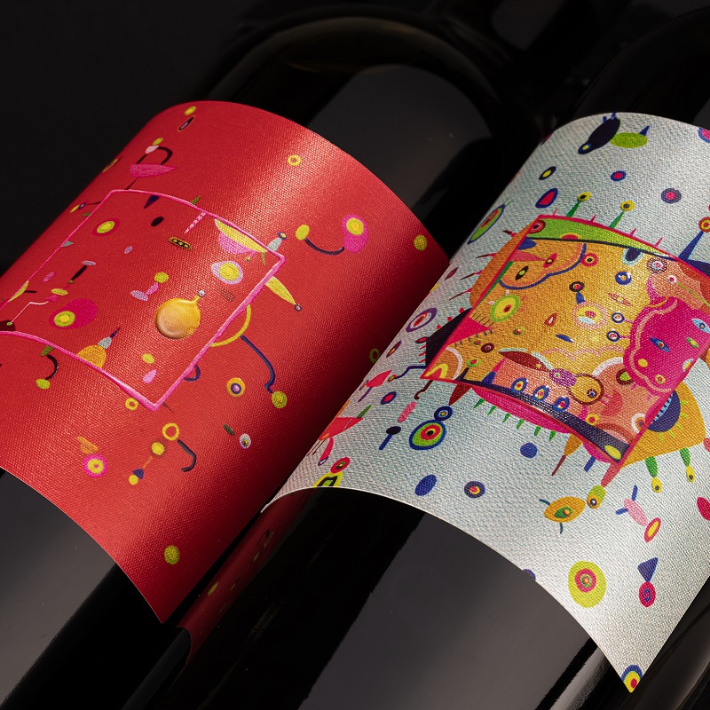 Zwei Weinflaschen stehen nebeneinander, jede mit einem farbenfrohen Etikett. Das rote Etikett zeigt bunte geometrische Muster, während das graue Etikett lebendige, verspielte Designs enthält.