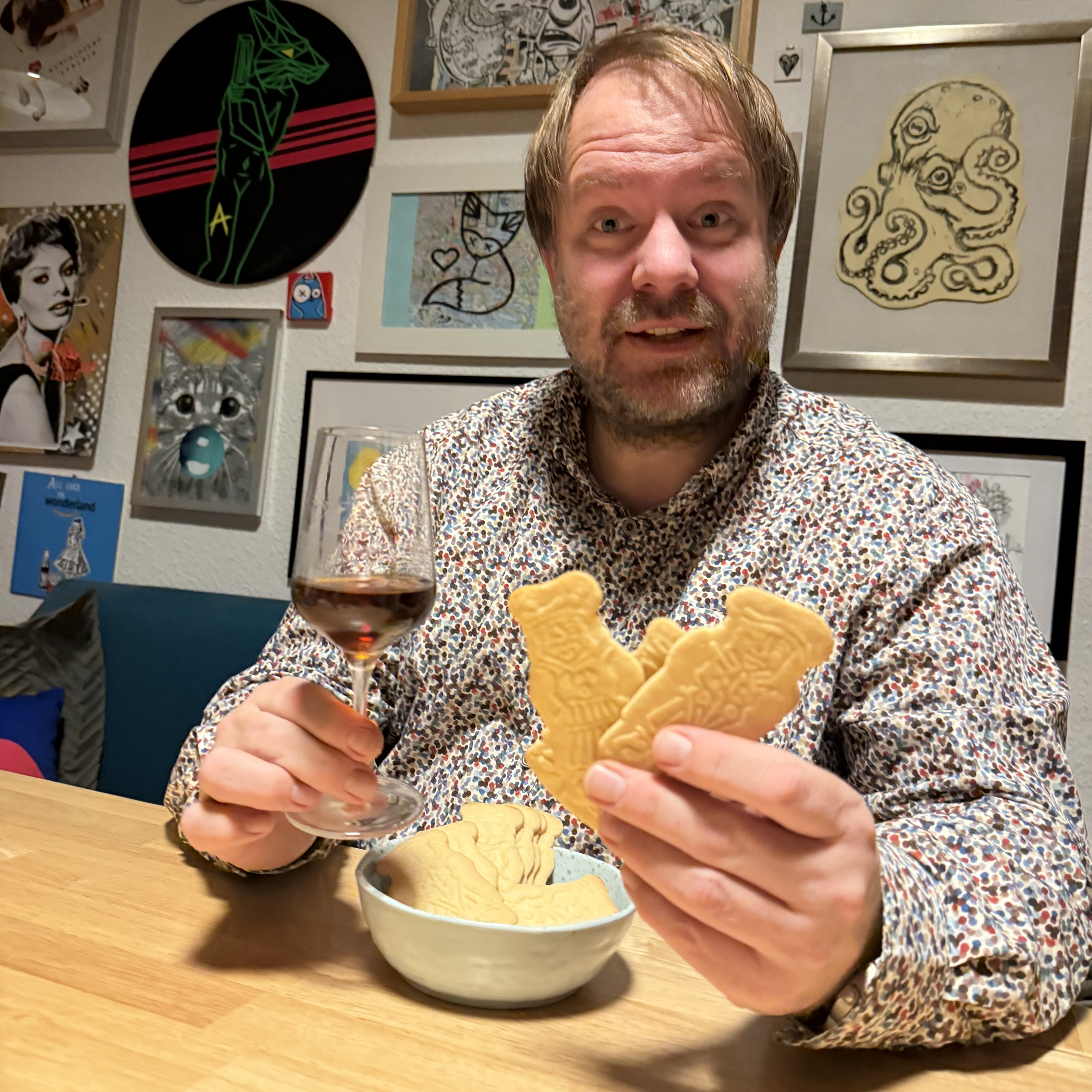 Eine Person mit einem bunten Hemd hält in der einen Hand ein Glas und in der anderen Hand mehrere gebratene Kekse. Im Hintergrund sind Kunstwerke an der Wand zu sehen. Die Szene spielt sich an einem Holztisch ab.