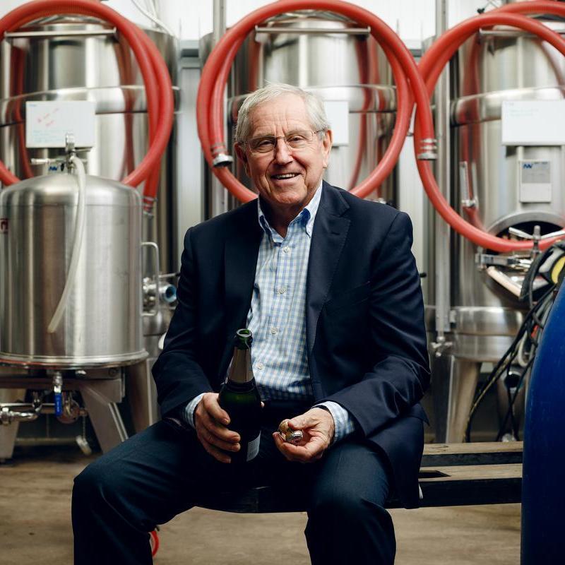 Ein älterer Mann sitzt auf einer Bank in einer Produktionsumgebung, hält eine Champagnerflasche in der Hand und lächelt. Im Hintergrund sind rostfreie Stahltanks und rote Schläuche zu sehen.