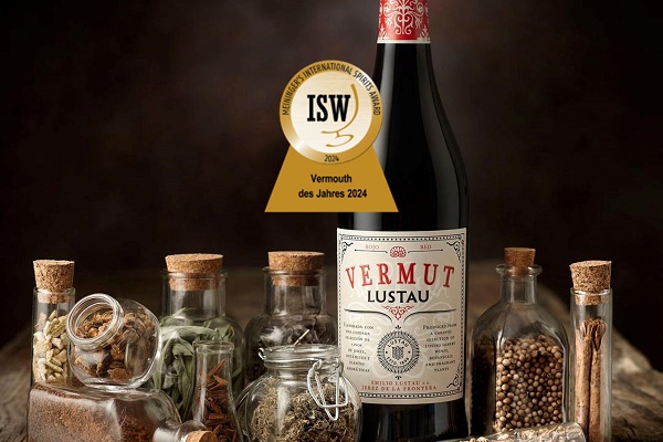 2_22.08._Lustau Eine Flasche Vermut steht im Mittelpunkt, umgeben von verschiedenen Gewürzen und Kräutern in Glasbehältern. Ein Preisetikett zeigt -Vermouth des Jahres 2024-. Der Hintergrund ist dunkel und schattig.